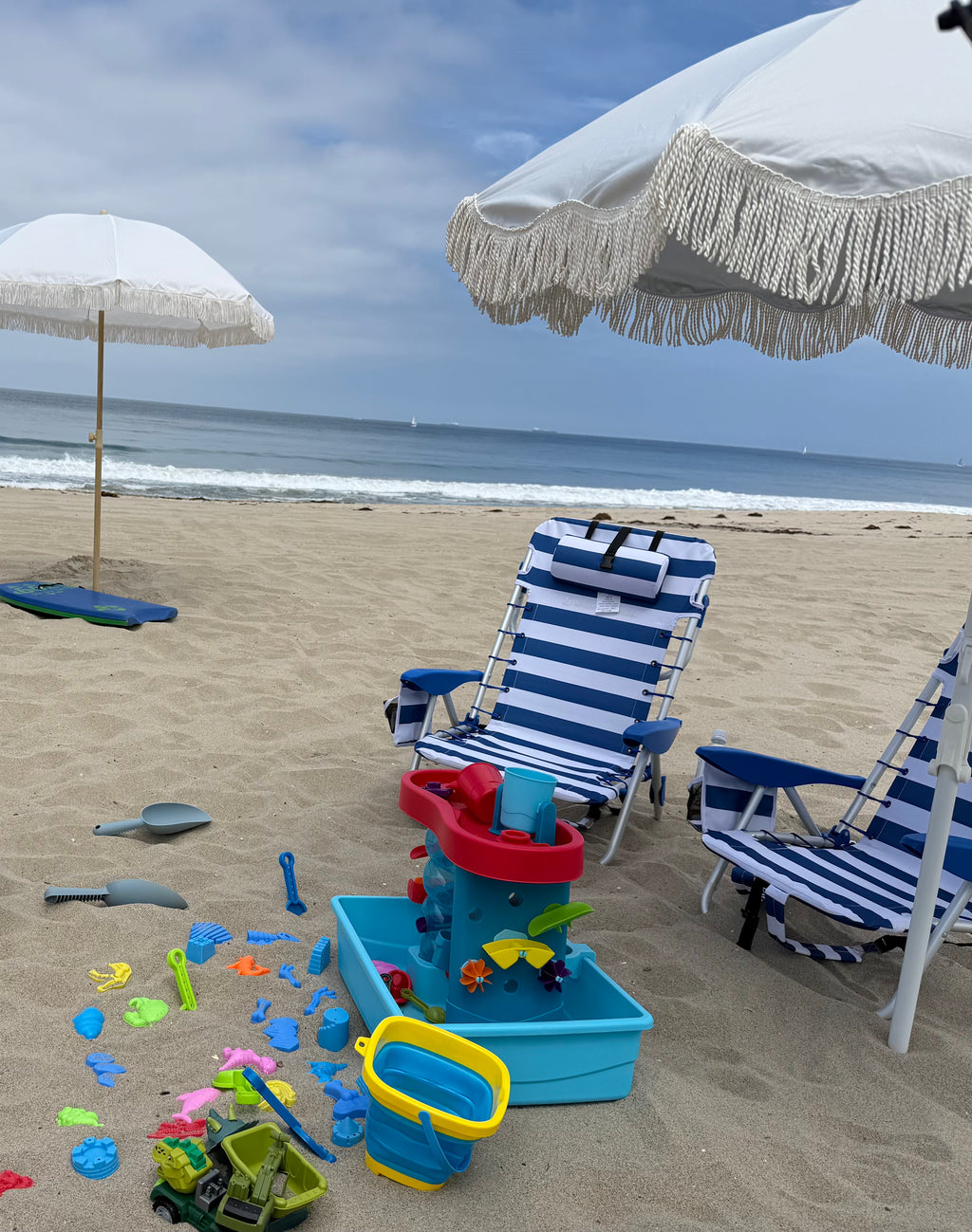 A beach scene with two blue-and-white striped lounge chairs under white umbrellas, a small blue sandbox with colorful toys, and the ocean in the background.