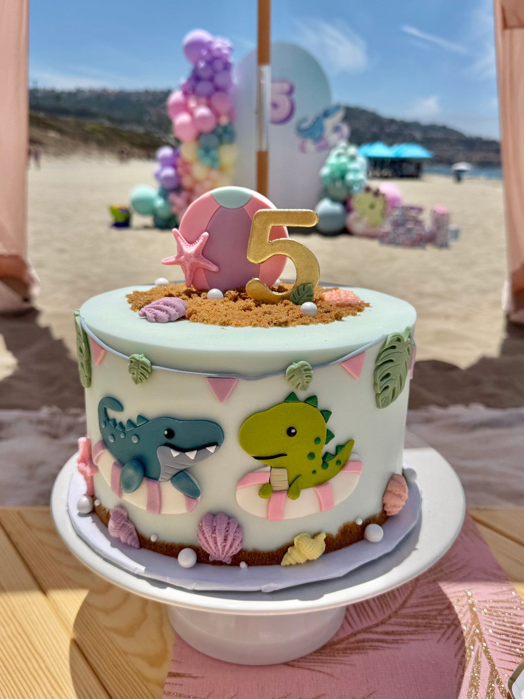 A round birthday cake decorated with pastel blue icing, dinosaur figures, seashells, sand-like topping, and a gold number 5 topper, displayed on a white cake stand with a beach-themed event background.