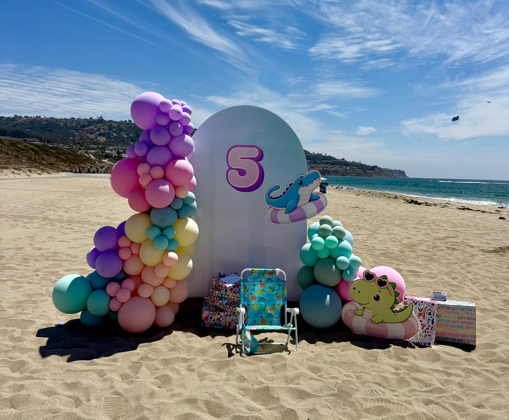 A beach-themed 5th birthday party setup on sandy shore with a large white arch backdrop featuring a colorful number 5 and mermaid illustration, surrounded by pastel balloon garlands, a small beach chair, gift bags, and a dinosaur float. No people or animals are present.