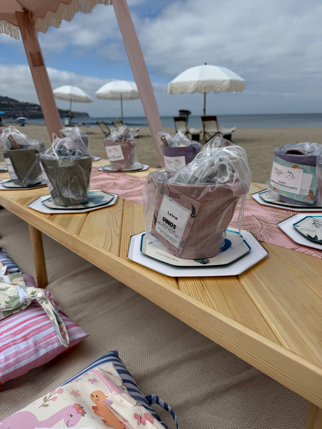 A wooden beach table set up on a sandy beach under a pink canopy, holding several wrapped bath or body products in clear plastic bags with paper labels. White beach umbrellas, lounge chairs, and the ocean are visible in the background.