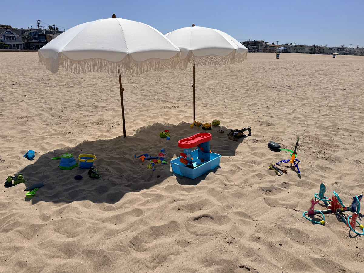 Two white beach umbrellas on a sandy beach with children's toys scattered around, including a blue sand castle, shovel, and other play items.